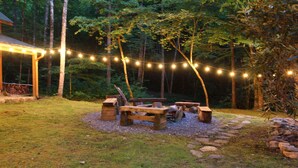 A wooden deck with a fire pit and benches, surrounded by a stone border and string lights. - Creek Front Mountain Cabin with Jacuzzi. (Andrews)
