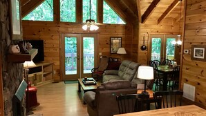 A cozy cabin interior with a fireplace, a sofa, a dining table, and large windows offering a view of the forest. - Creek Front Mountain Cabin with Jacuzzi. (Andrews)