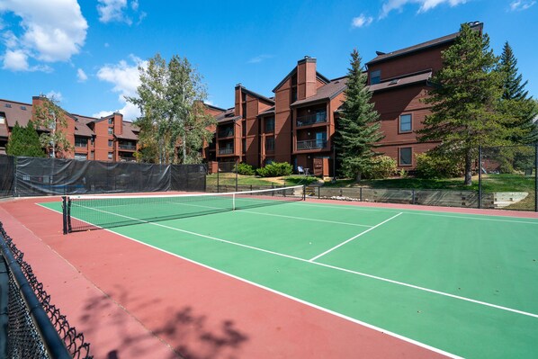 A tennis court with a net, surrounded by a fence, and adjacent to a building with multiple balconies and a brick facade. - Ski Week Basecamp | Ski Shuttle + Fireplace (Steamboat Springs)
