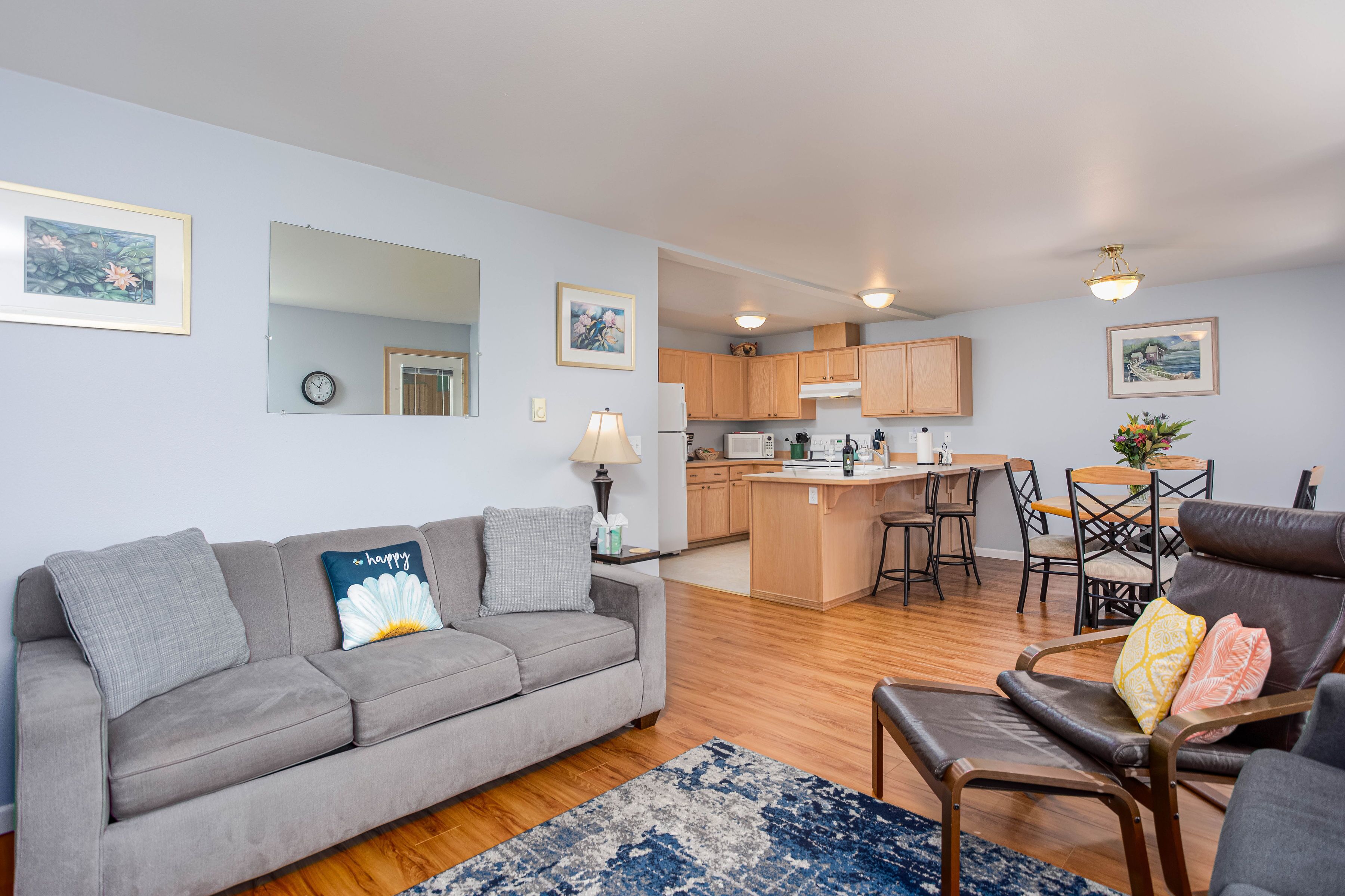 Una sala de estar moderna con un sofá gris, una mesa de centro y un sillón de cuero. Se ve una cocina con armarios de madera y un comedor con mesa y sillas al fondo.