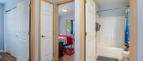 A hallway with a bedroom and a bathroom. The bedroom has a bed with a red blanket and a dresser. The bathroom has a bathtub and a toilet.
