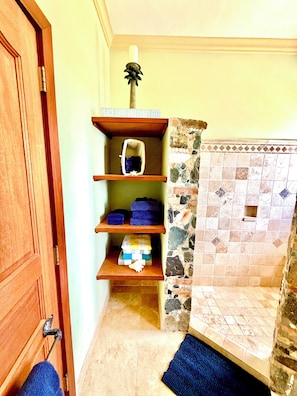A bathroom with a wooden door, a shelf with folded towels, a stone-tiled shower area, and a blue bath mat.