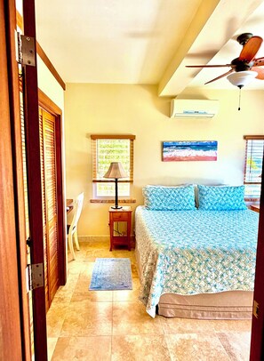 A bedroom with a bed, a ceiling fan, a window with blinds, a nightstand, a lamp, a chair, and a rug. - COUPLES RETREAT, PANORAMIC OCEAN VIEWS, FREE KAYAKS & PADDLE BOARDS (St. John)