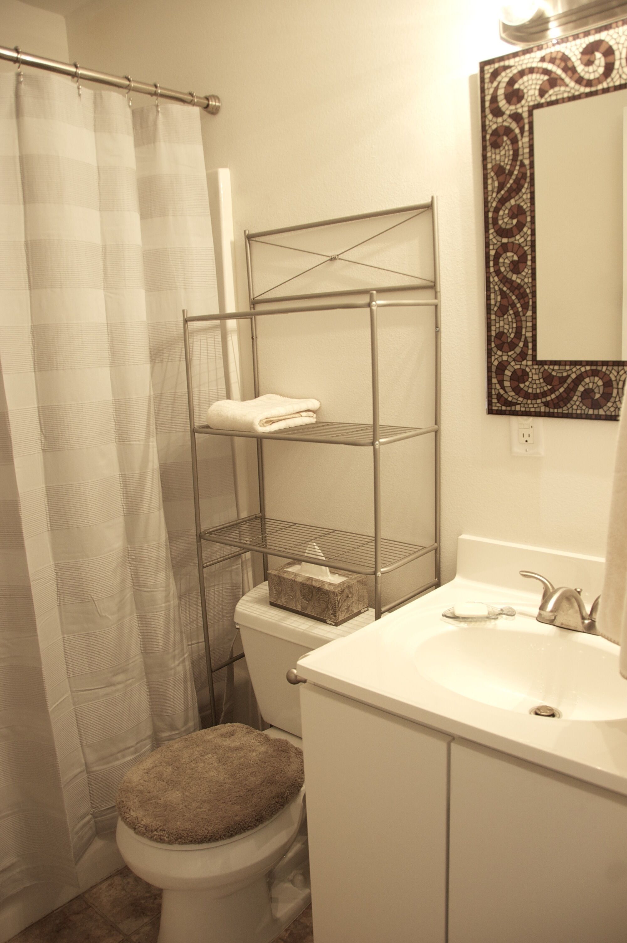 A bathroom with a shower curtain, a toilet with a beige seat, a metal shelf unit with a folded towel, a sink with a faucet, and a decorative mirror with a mosaic frame.