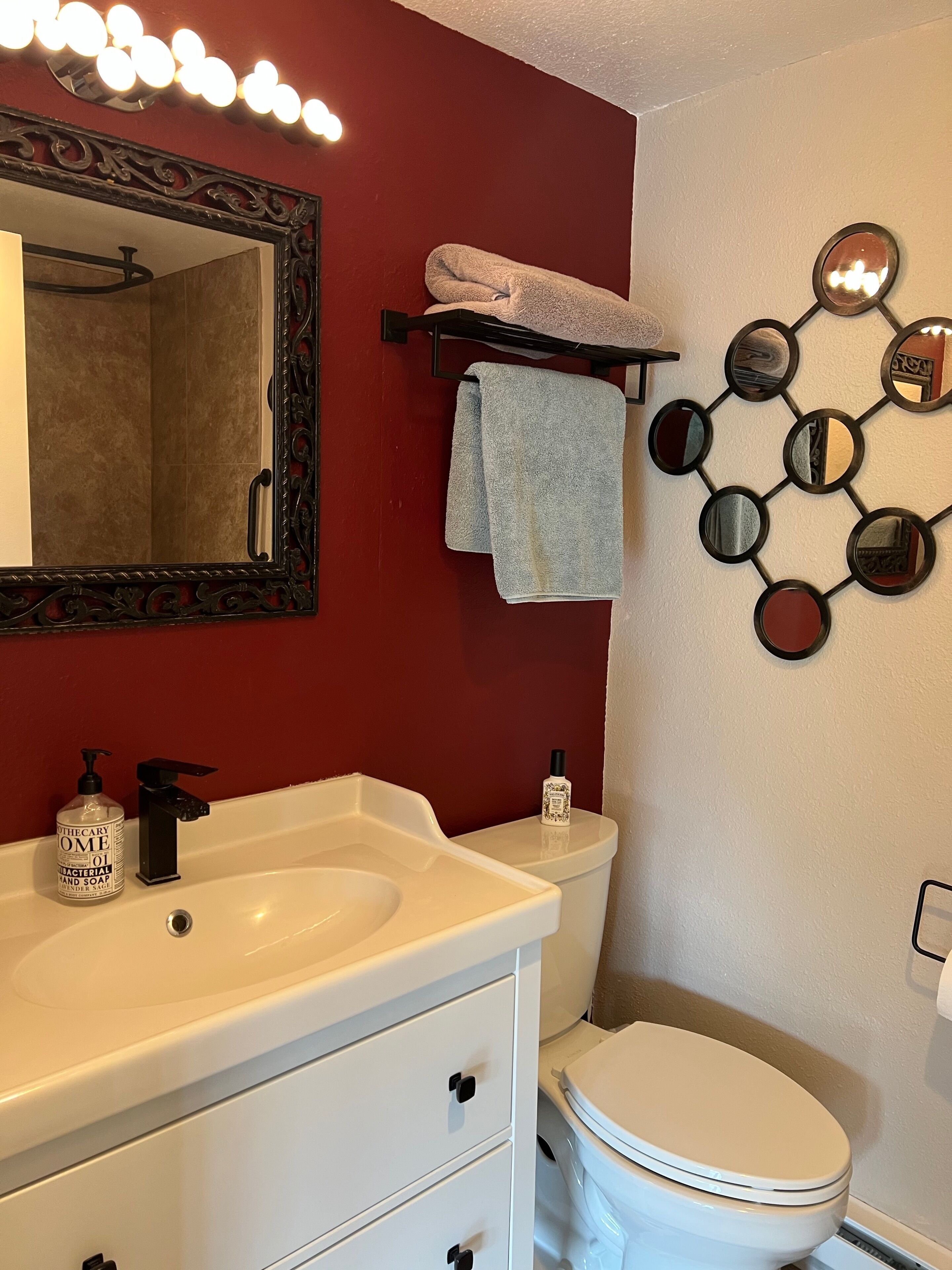 Une salle de bain avec un lavabo blanc, un robinet noir et un miroir surmonté de lumières. On y trouve également des toilettes blanches et un porte-serviettes avec une serviette.