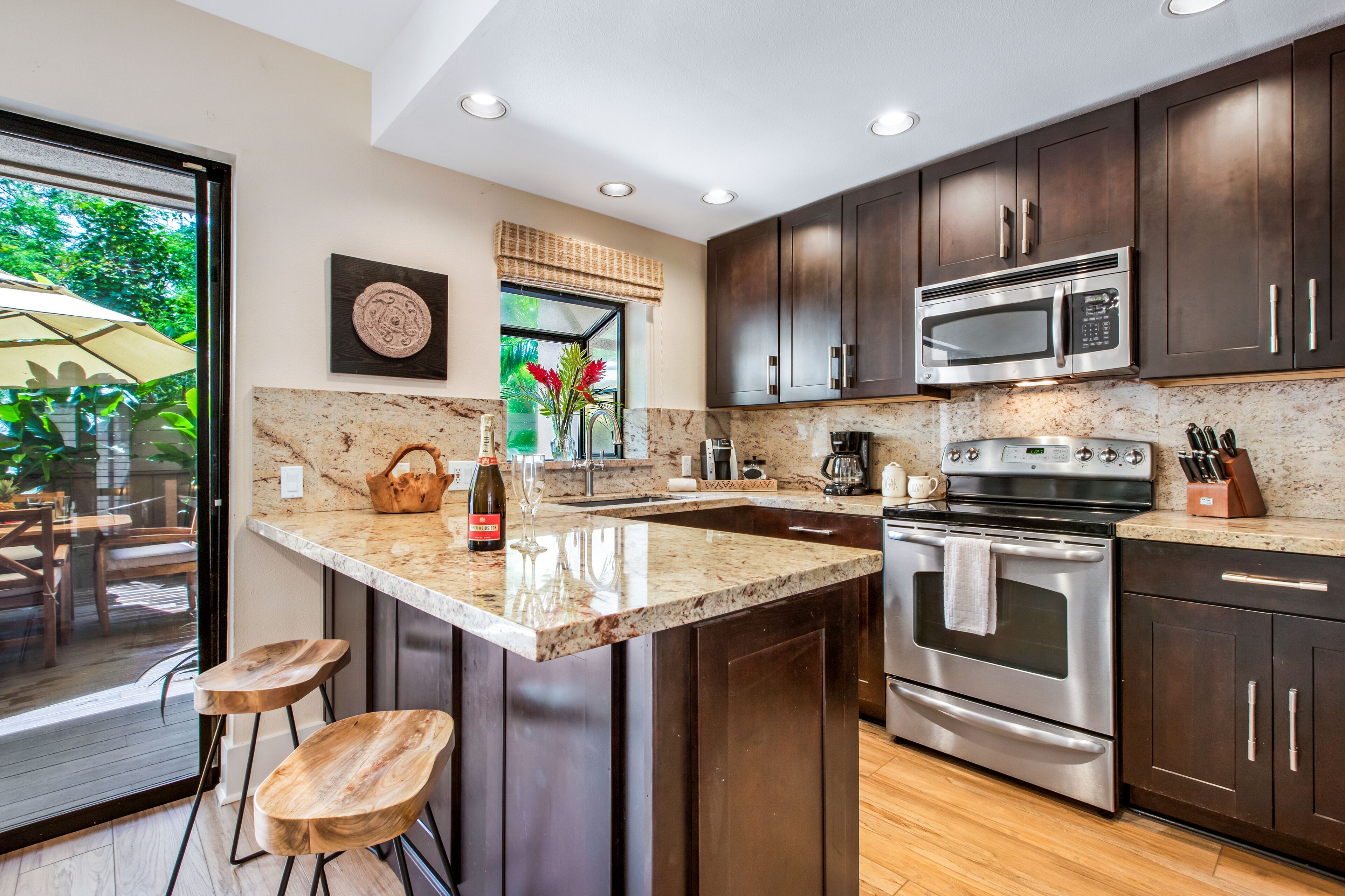 A modern kitchen with dark wood cabinets, stainless steel appliances, a large island with stools, and a view to the outside.