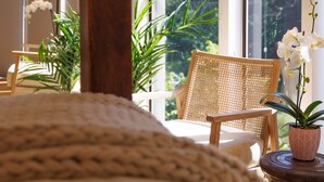Een slaapkamer met een groot raam, een houten bed, een rieten stoel en een nachtkastje met een plant.