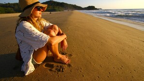 Eine Person sitzt an einem sandigen Strand mit Blick auf den Ozean und ferne Hügel.