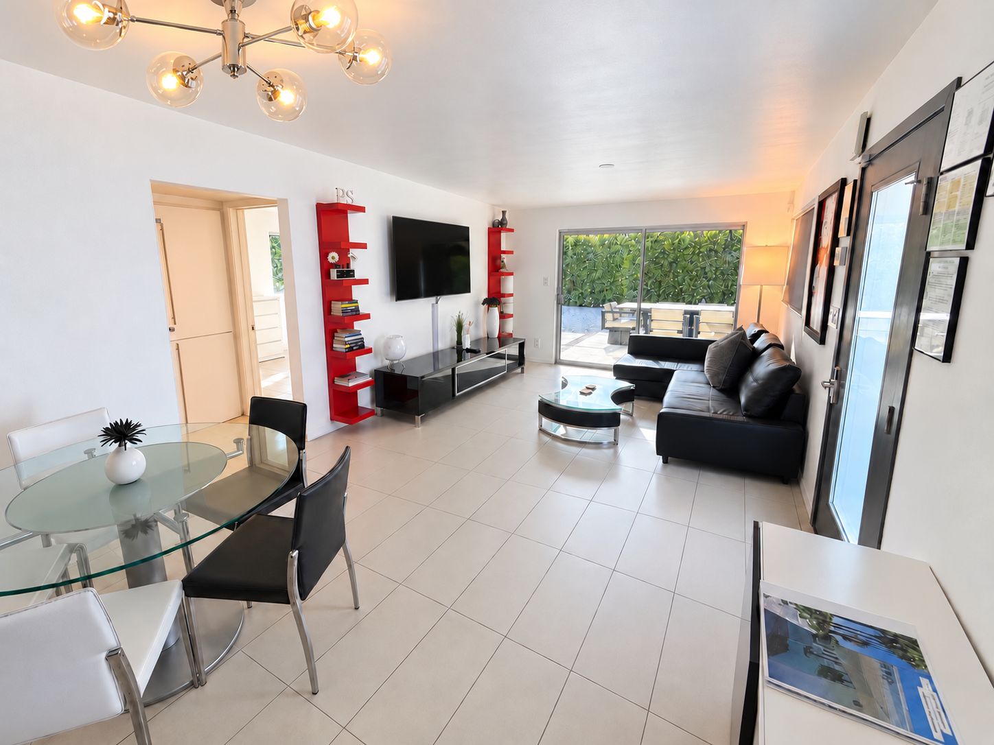 A modern living room with a glass dining table, black leather sofa, and a flat-screen TV mounted on the wall.