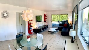 A modern living room with a glass dining table, black and white sofas, a TV, and a large window. - Iconic Mid-Century Alexander Home in Palm Springs w/ Pool & Spa (Palm Springs)