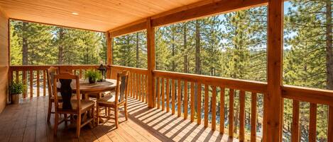 A wooden deck with a table and chairs, surrounded by a railing, overlooking a forested area.