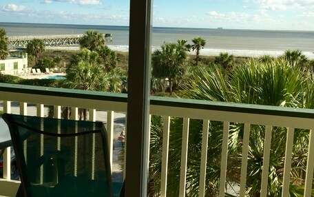 A balcony with a view of the beach, palm trees, and a pier in the distance.
