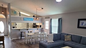 Una sala de estar moderna con un sofá azul, un comedor con sillas blancas y una cocina con microondas y refrigerador.
