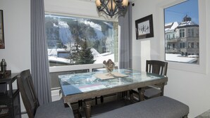 A dining area with a glass table, grey upholstered chairs, a bench, a chandelier, and a view of a snowy mountain landscape through large windows.