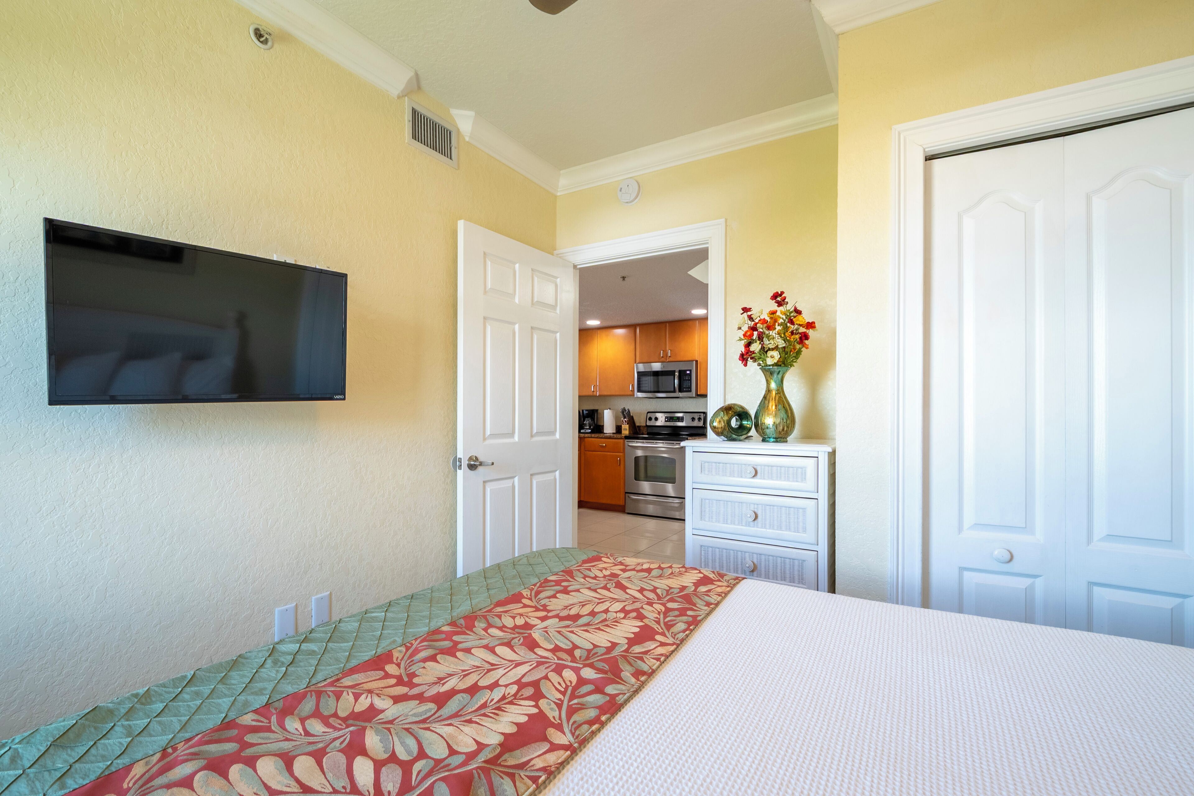 Un dormitorio con cama, televisor, puerta y vista a la cocina.