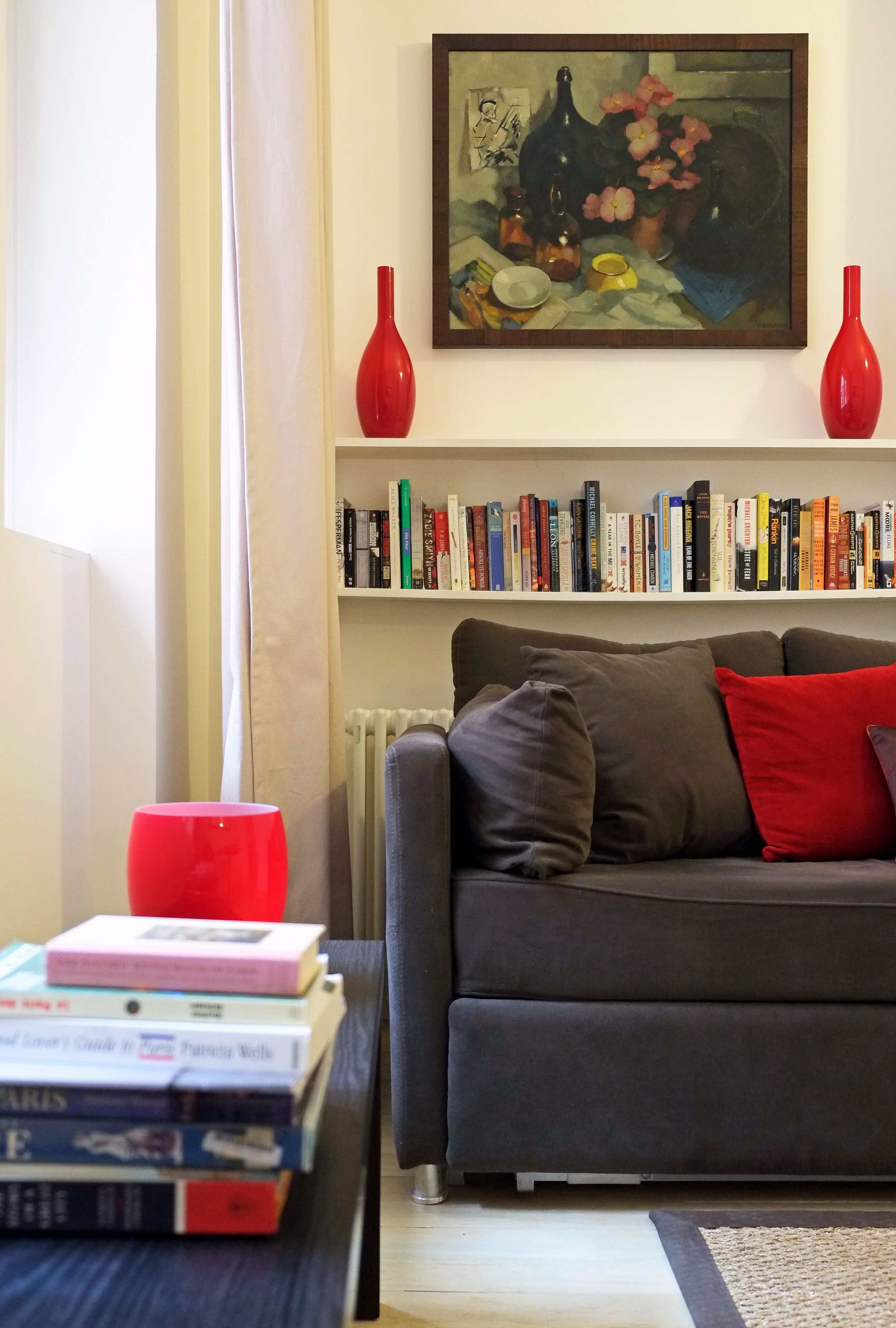 Un salon avec un canapé gris, des coussins rouges et bruns, un vase rouge et une bibliothèque remplie de livres.