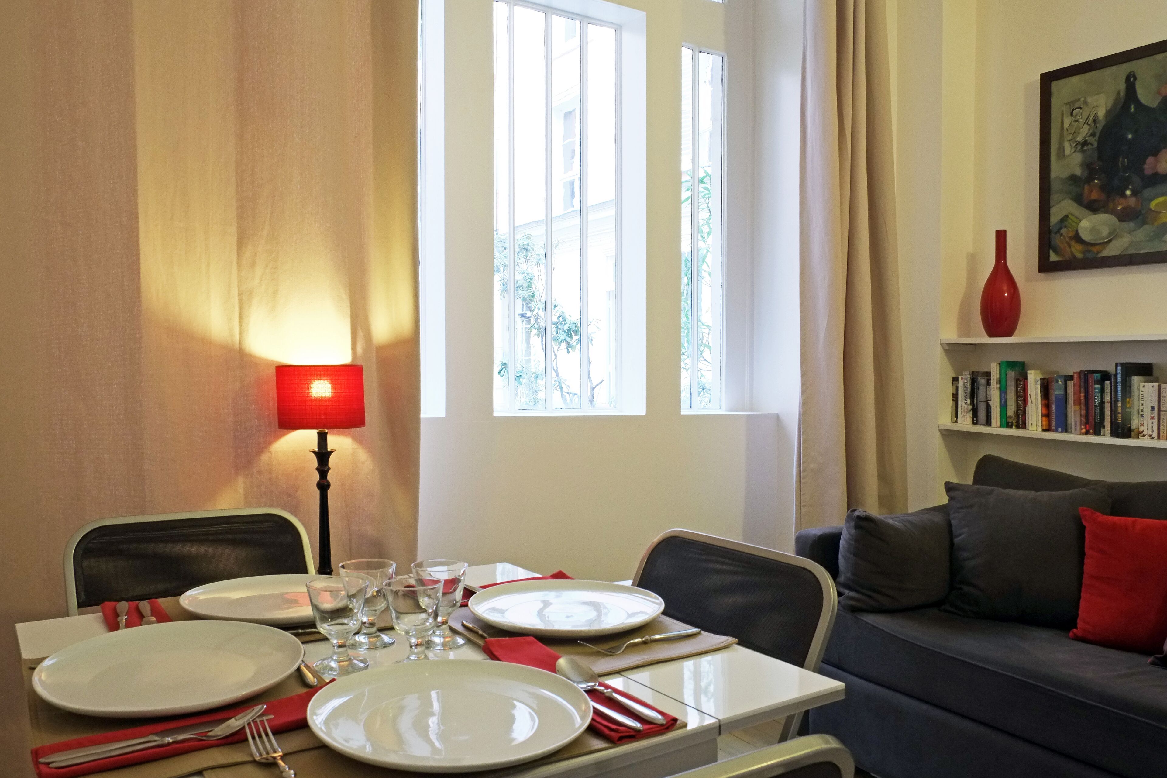 A dining area with a table set for two, a red lamp, and a bookshelf with books.