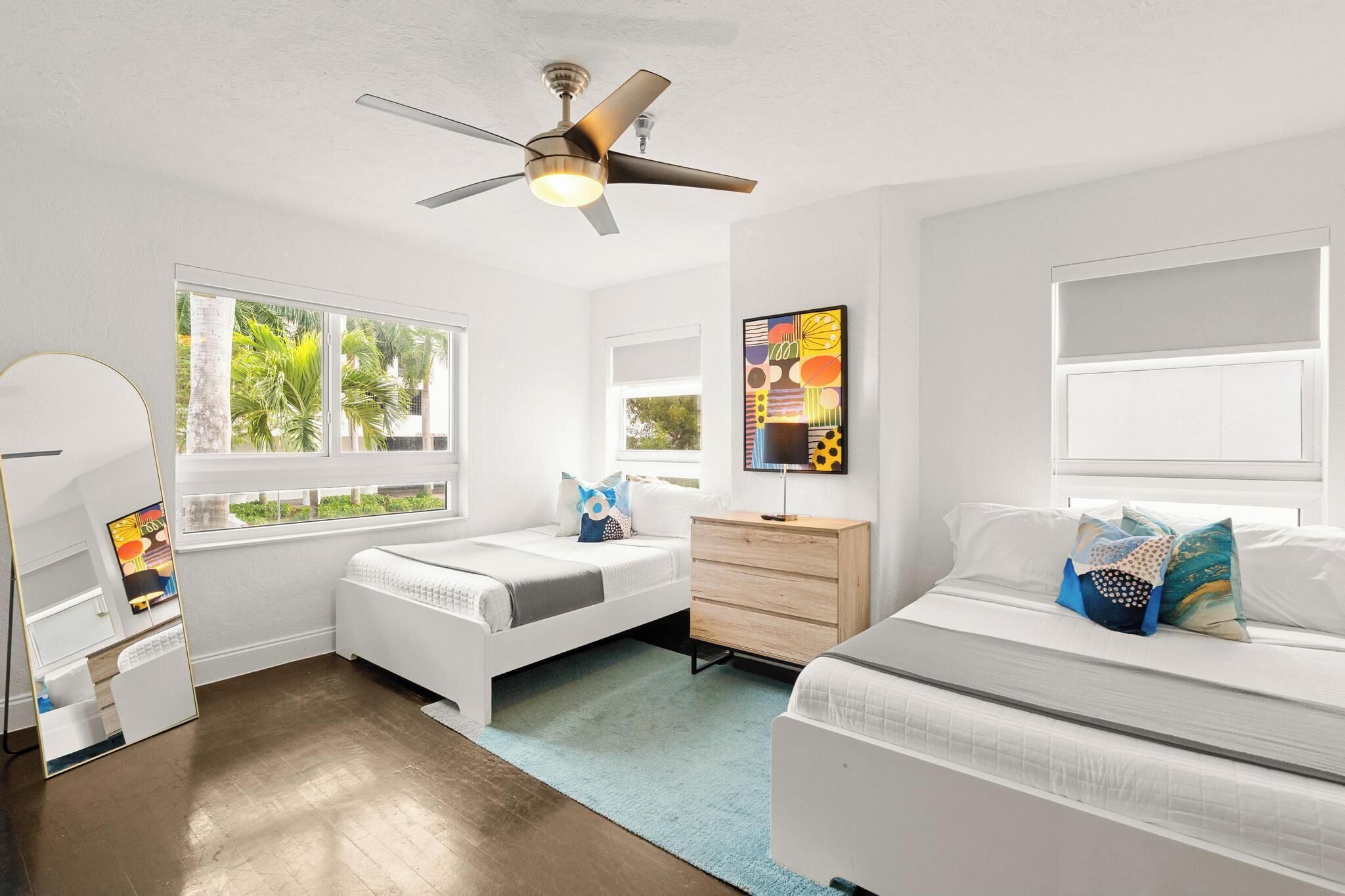 Une chambre moderne avec deux lits, un ventilateur de plafond, une commode et une grande fenêtre donnant sur des palmiers.
