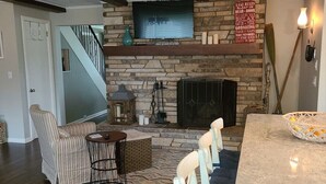 A cozy living room with a stone fireplace, a television mounted above it, and a dining area with a bar. - FAMILY FRIENDLY COTTAGE SLEEPS 12 !!! BEAUTIFULLY UPDATED (Petoskey)