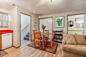 A living room with a brown sofa, a dining table with chairs, a bookshelf, and a staircase leading to another room.