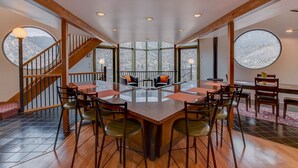 Un'ampia sala da pranzo con soffitto alto, ampie finestre che offrono vista sulle montagne e una scala in legno che conduce a un piano superiore.