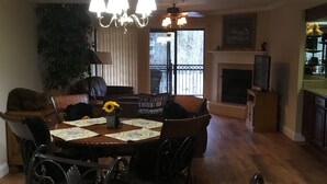 A dining area with a round table, chairs, a chandelier, a fireplace, and a large window with a view of the outdoors.