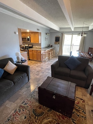 A living room with a sofa, a loveseat, a coffee table, and a rug. There is a kitchen in the background with wooden cabinets and a dining area with a table and chairs.