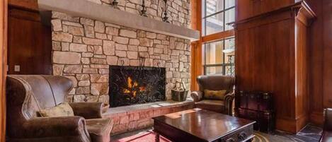 A stone fireplace with a fire, leather sofas, and a wooden coffee table in a room with wooden walls and large windows.