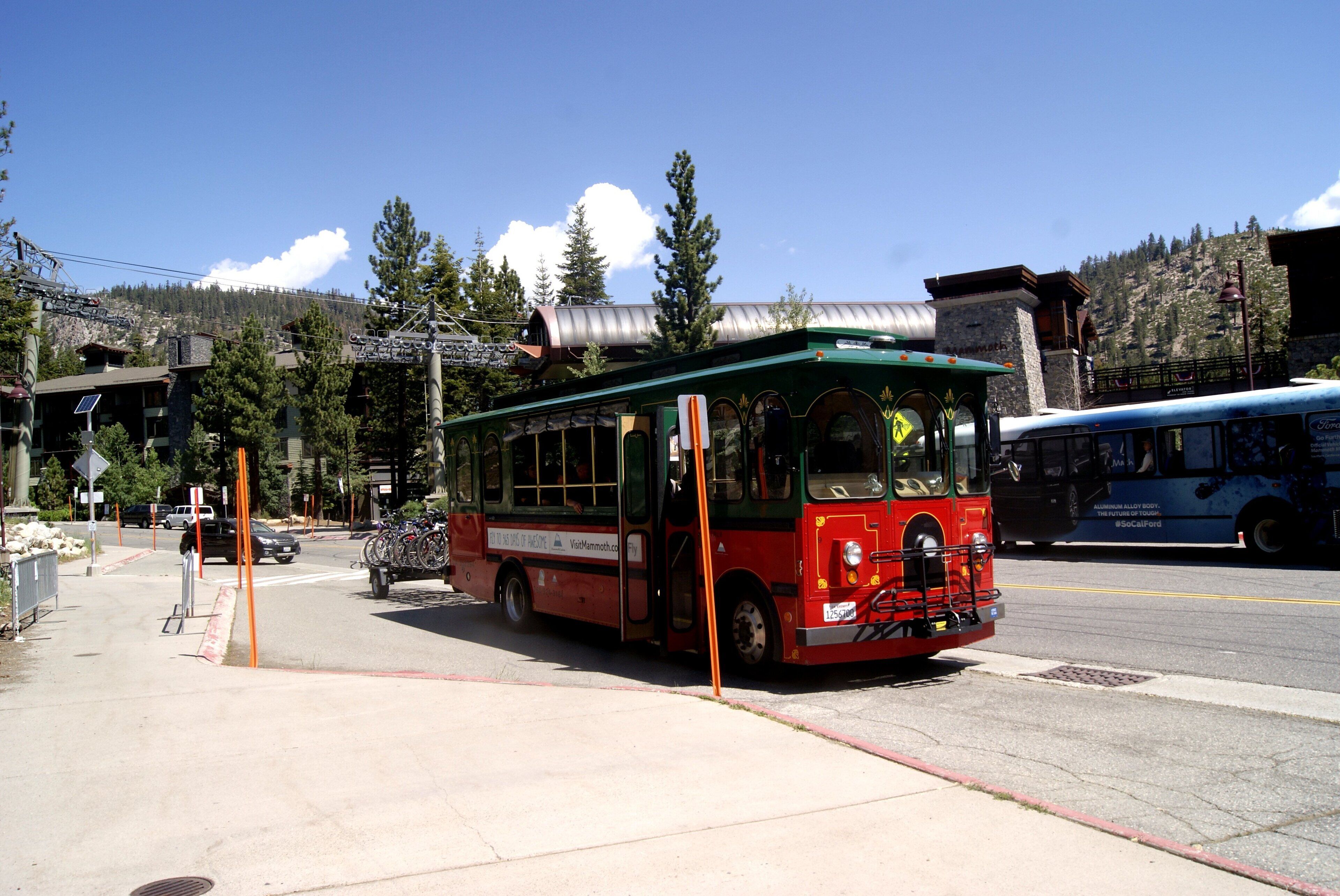 A red and green trolley bus parked at a bus stop with a mountainous backdrop.
