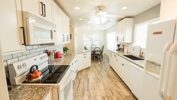 A kitchen with white cabinets, a granite countertop, a stove, a microwave, a ceiling fan, and a dining area with a table and chairs.