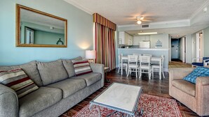 A living room with a sofa, coffee table, and striped curtains. A kitchen is visible in the background with white cabinets and a dining area.