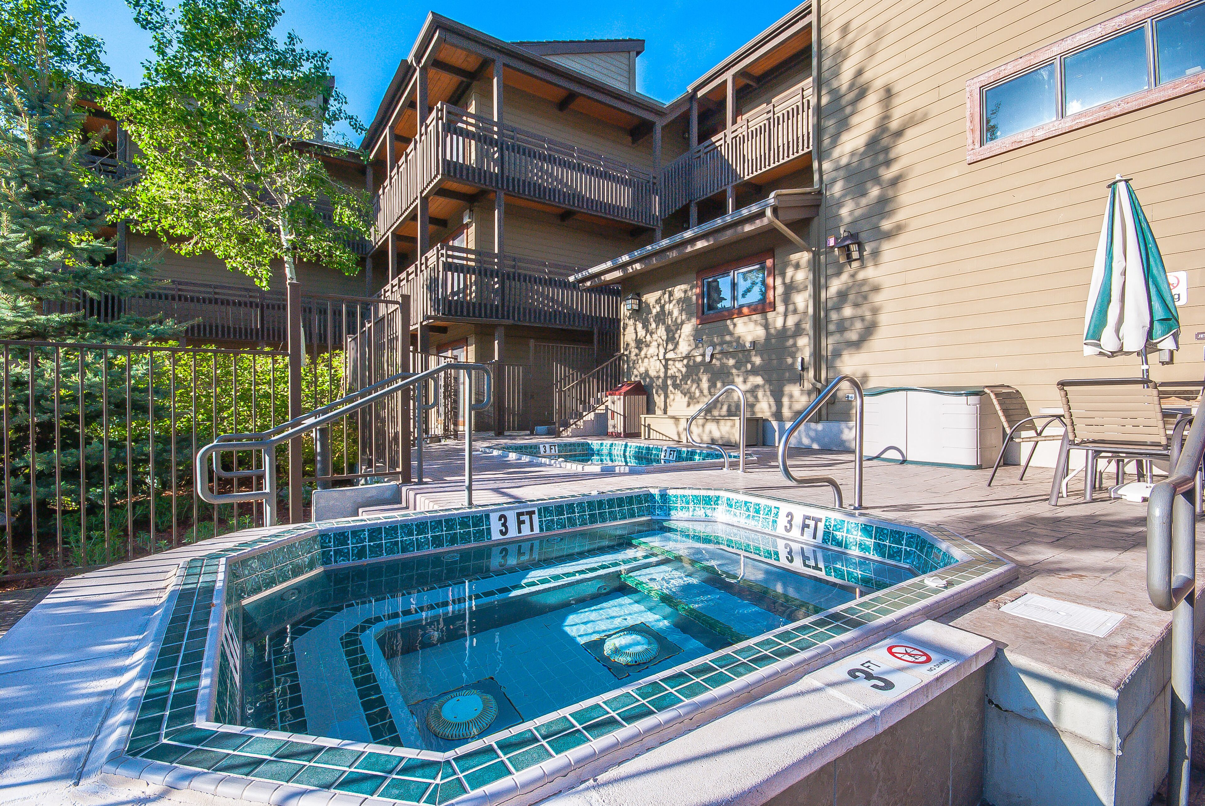A hot tub with a 3 FT depth, surrounded by a wooden deck and a poolside area with lounge chairs and an umbrella.