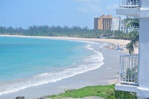 A beach view with a clear sky, a sandy shore, and a few people. There are buildings in the background and a white structure in the foreground. - Location,Location,Location "Casa Gelma" WATERFRONT  (Isla Verde)