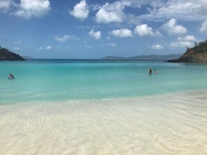 Ein Strand mit klarem türkisblauem Wasser, einem sandigen Ufer und Bergen in der Ferne.
