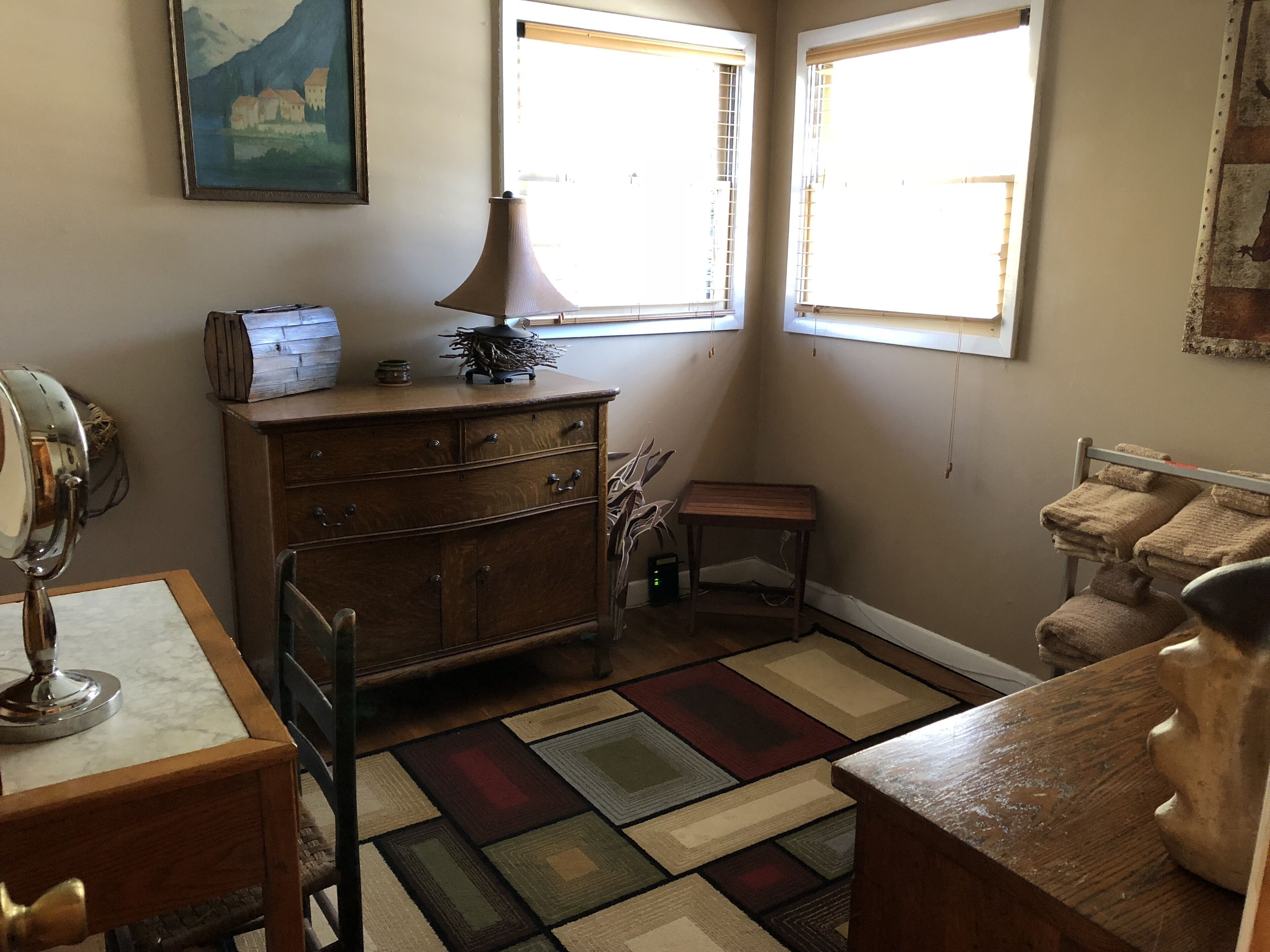 A cozy room with a wooden dresser, a small table, a chair, a lamp, and a painting on the wall.