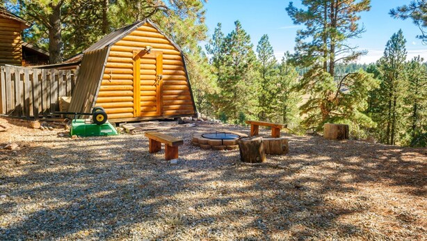 A log cabin with a sloped roof, surrounded by a gravel area with a fire pit, benches, and a green lawnmower. - Romantic Mountain Cabin | Near Bryce & Zion (Duck Creek Village)