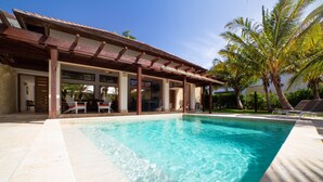 A swimming pool with a wooden deck and lounge chairs, surrounded by palm trees and a covered patio area.