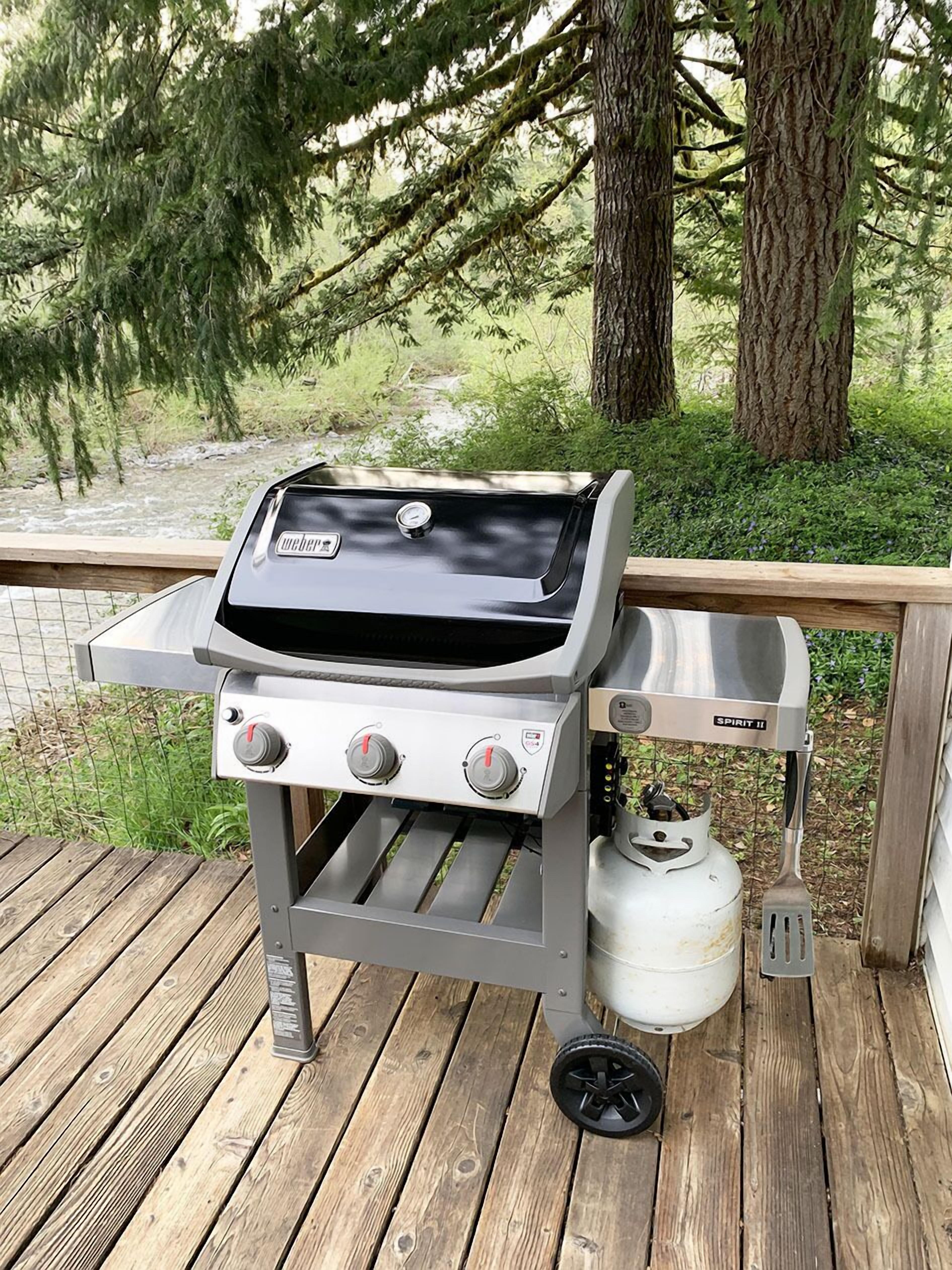 Una griglia Weber Spirit II portatile con bombola del gas e bruciatore laterale, posizionata su un pavimento in legno.