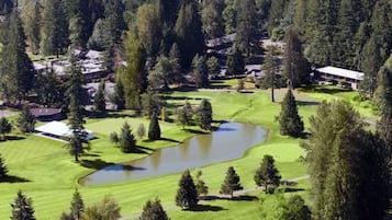 Surrounded by dense forest, this golf course features well-manicured grass and a variety of trees, including tall conifers, with a serene pond and distant resort buildings.