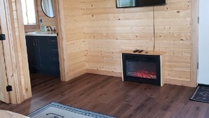 A wooden-paneled room with a fireplace, a mounted TV, and a bathroom with a mirror and sink. - Kia'a'yo Bear Cabin -- Modern Cabin -- 15 Min to Glacier East Entrance (Babb)