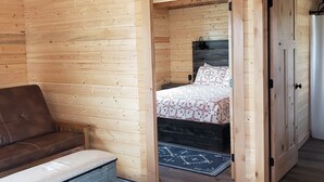 A wooden cabin interior with a sofa, a bed with patterned bedding, and a blue rug with geometric designs. - Kia'a'yo Bear Cabin -- Modern Cabin -- 15 Min to Glacier East Entrance (Babb)