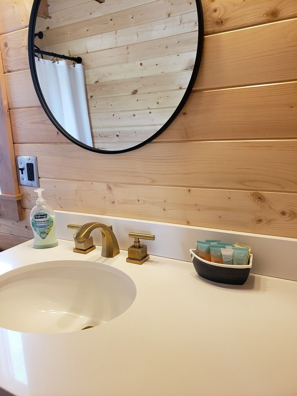 Een badkamer met een houten wand, een ronde spiegel, een witte wastafel, een gouden kraan en een zeepdispenser.