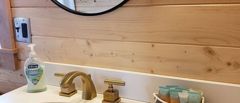 Un baño con pared de madera, un espejo redondo, un lavabo blanco, un grifo dorado y un dispensador de jabón.
