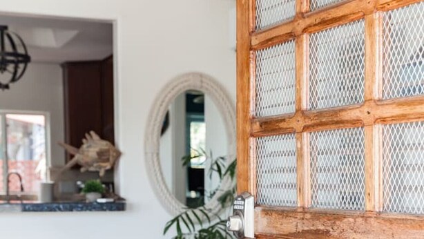 A wooden door with a metal mesh panel, a potted plant, and two metal stools in a room with wooden flooring and a round mirror on the wall.