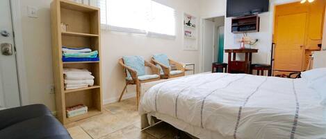 Habitación de hotel con una cama, dos sillas, un estante de madera con toallas, un televisor y una puerta.