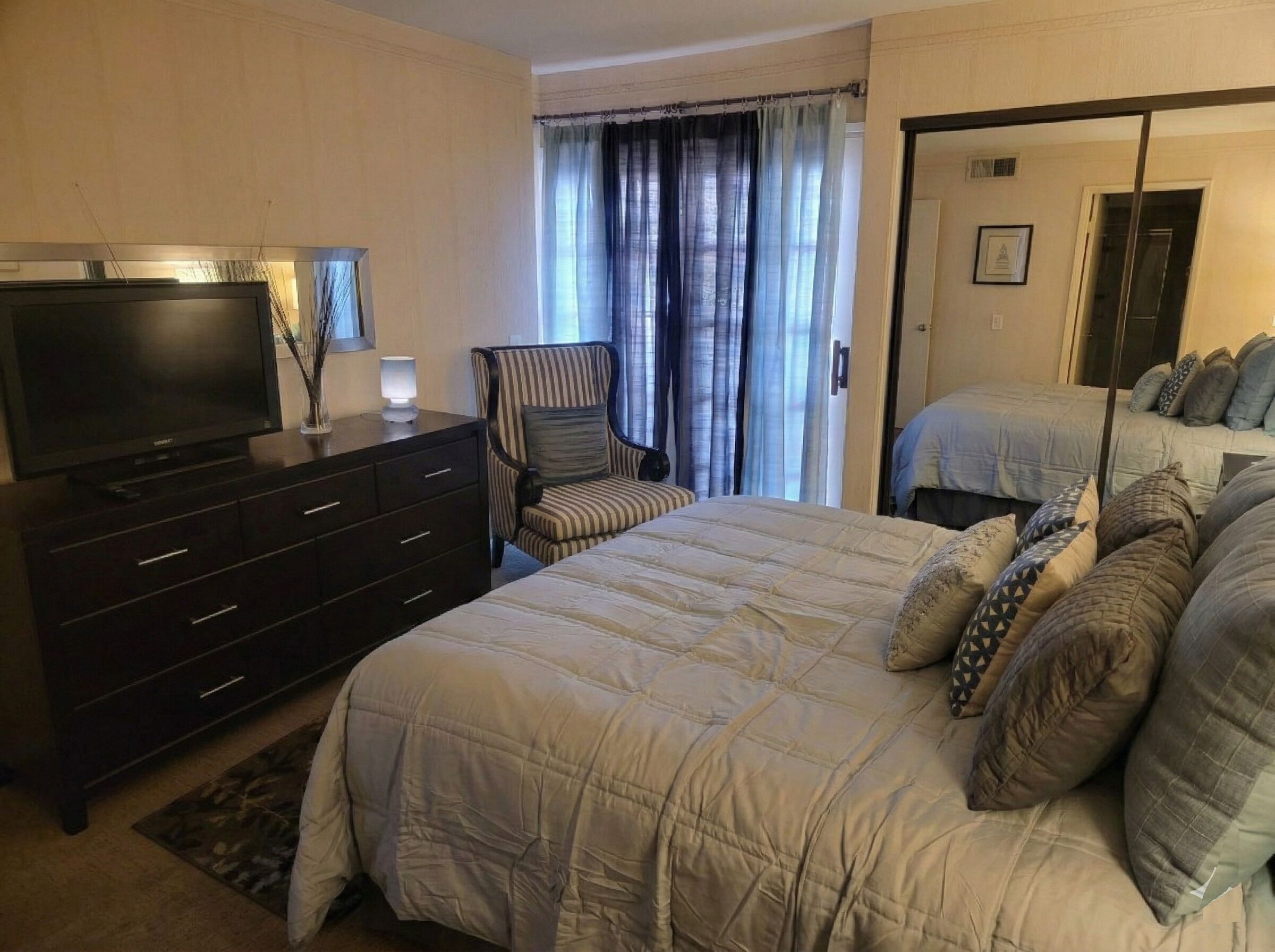 Une chambre d’hôtel avec un grand lit, un téléviseur posé sur une commode, une chaise et une porte donnant sur une autre pièce.