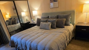 A neatly made bed with a striped duvet, a bedside table with a lamp, and a mirror on the wall. - Heart of Palm Springs (Palm Springs)