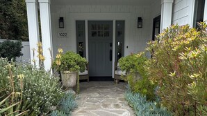 A stone pathway leading to a house with a grey door, numbered 1022, flanked by potted plants and shrubs.