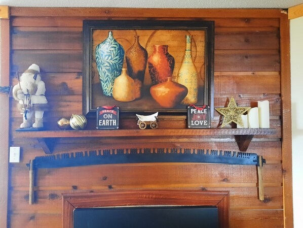 A wooden mantelpiece with a framed artwork of vases, a peace sign, candles, and a small figurine.