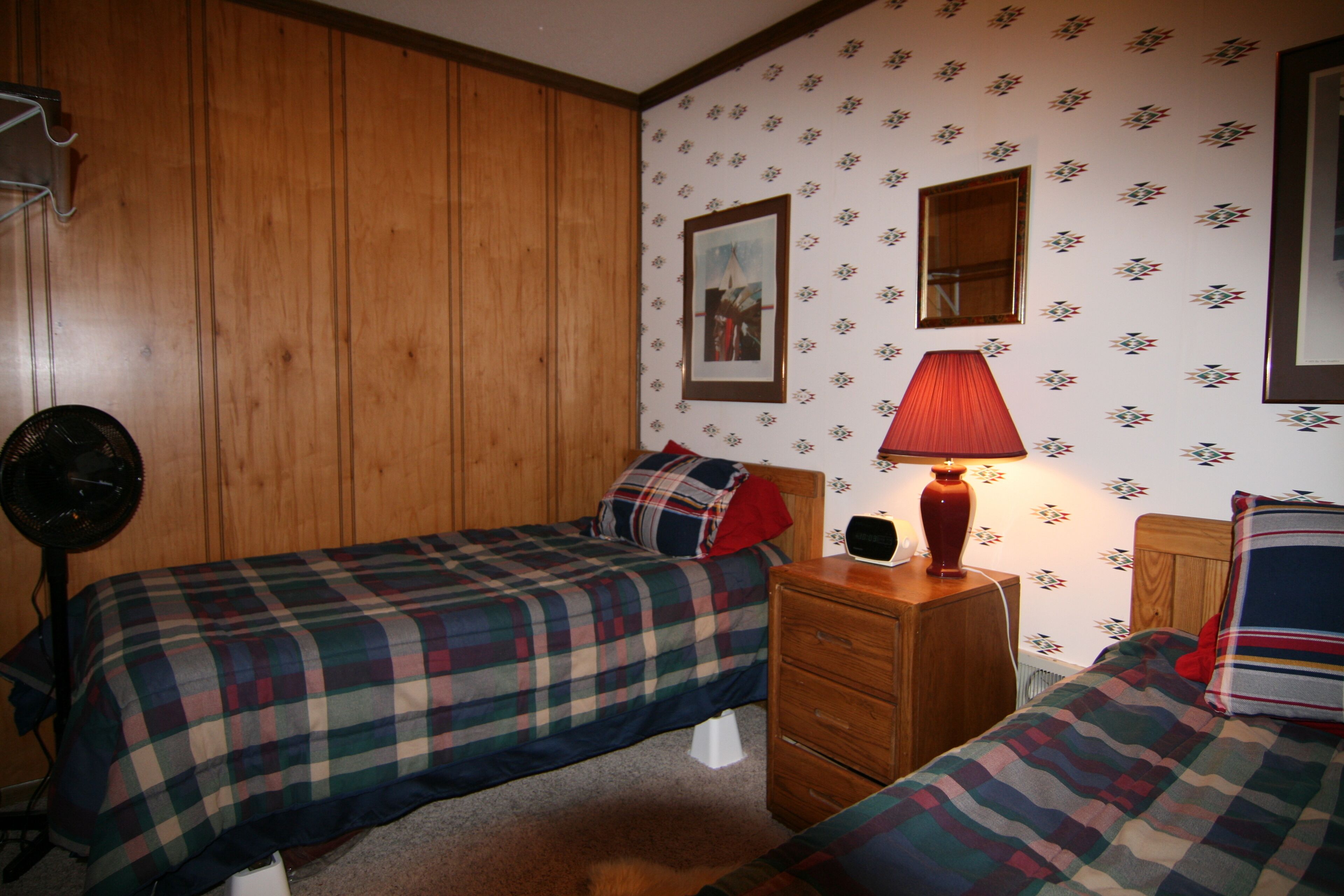 A room with two beds, wooden paneling, a fan, a lamp, and a framed picture on the wall.